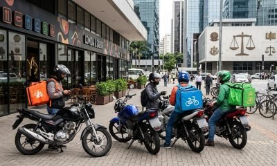 Entregadores da 99Food e outros apps de delivery em frente a um restaurante, ilustrando a investigação do Cade por abuso de posição e práticas anticompetitivas.