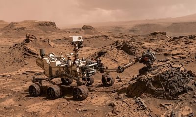 O Rover Curiosity em Marte, com seu braço robótico examinando rochas no Boxwork Terrain, sob um céu avermelhado e empoeirado, destacando a exploração do Boxwork.